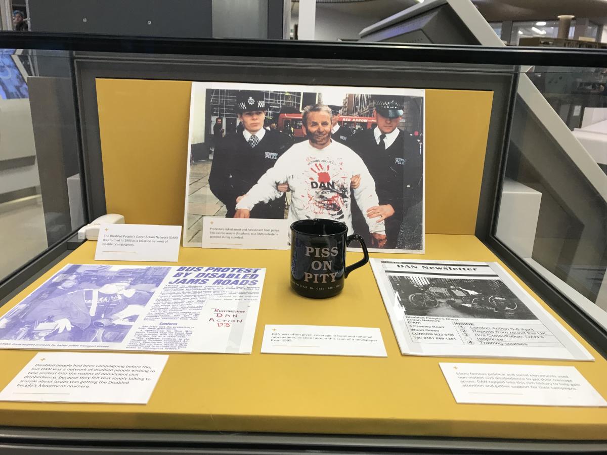 Glass exhibition case filled with photos and documents on a yellow background. The central photo is of a person in a white DAN sweatshirt smeared with black paint as they are taken away by two police officers. In front of the photo is a black mug printed with 'Piss on Pity'. Other documents include a DAN newsletter and a newspaper clipping that reads 'Bus protest by disabled jams roads'.