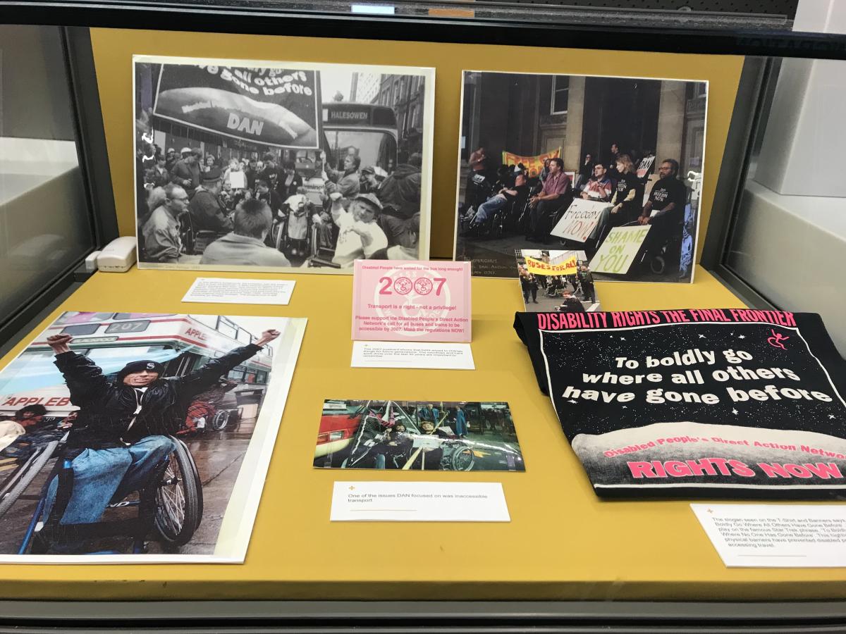 Glass exhibition case with DAN protest photos and a t-shirt on a yellow background. Back left photo is of many people at a transport protest in front of a bus, holding a banner. Back right photo is of 10 people protesting in front of a building in Birmingham holding signs that read 'Shame on You' and 'Freedom Now'. Front left photo is a person in a wheelchair in front of a bus with both arms in the air in triumph. Front right is a black t-shirt with a starry sky image reading 'Disability Rights, the final frontier. To Boldly go where all others have gone before. Disabled People's Direct Action Network, Rights Now.'