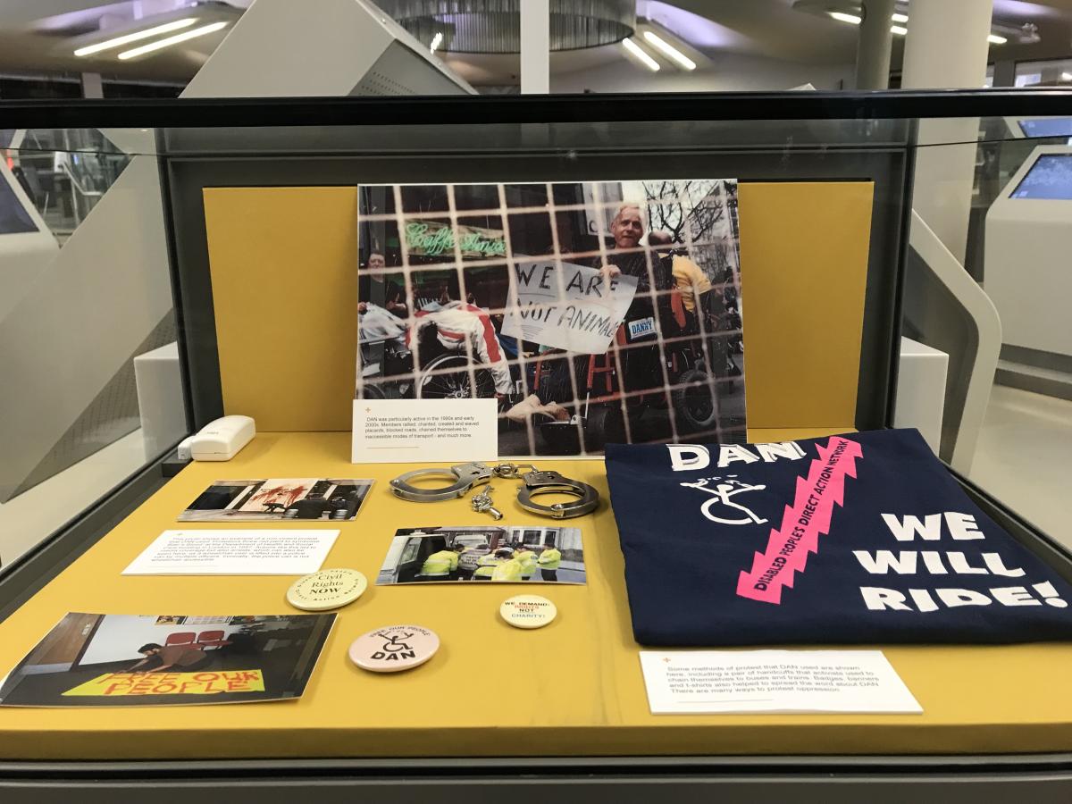 Glass exhibition case with DAN protest objects and photos on a yellow background. Central photo is a wheelchair user holding a sign 'We are not animals' with a square mesh in front of the camera lens. In front of this is a pair of metal handcuffs and several circular badges. On the left are several smaller photos of a building splattered with red paint, a wheelchair user lifted into a van by police officers and a person making a sign that reads 'Free our people'. On the right is a navy blue t-shirt printed with 'DAN We will ride' and a wheelchair user symbol.