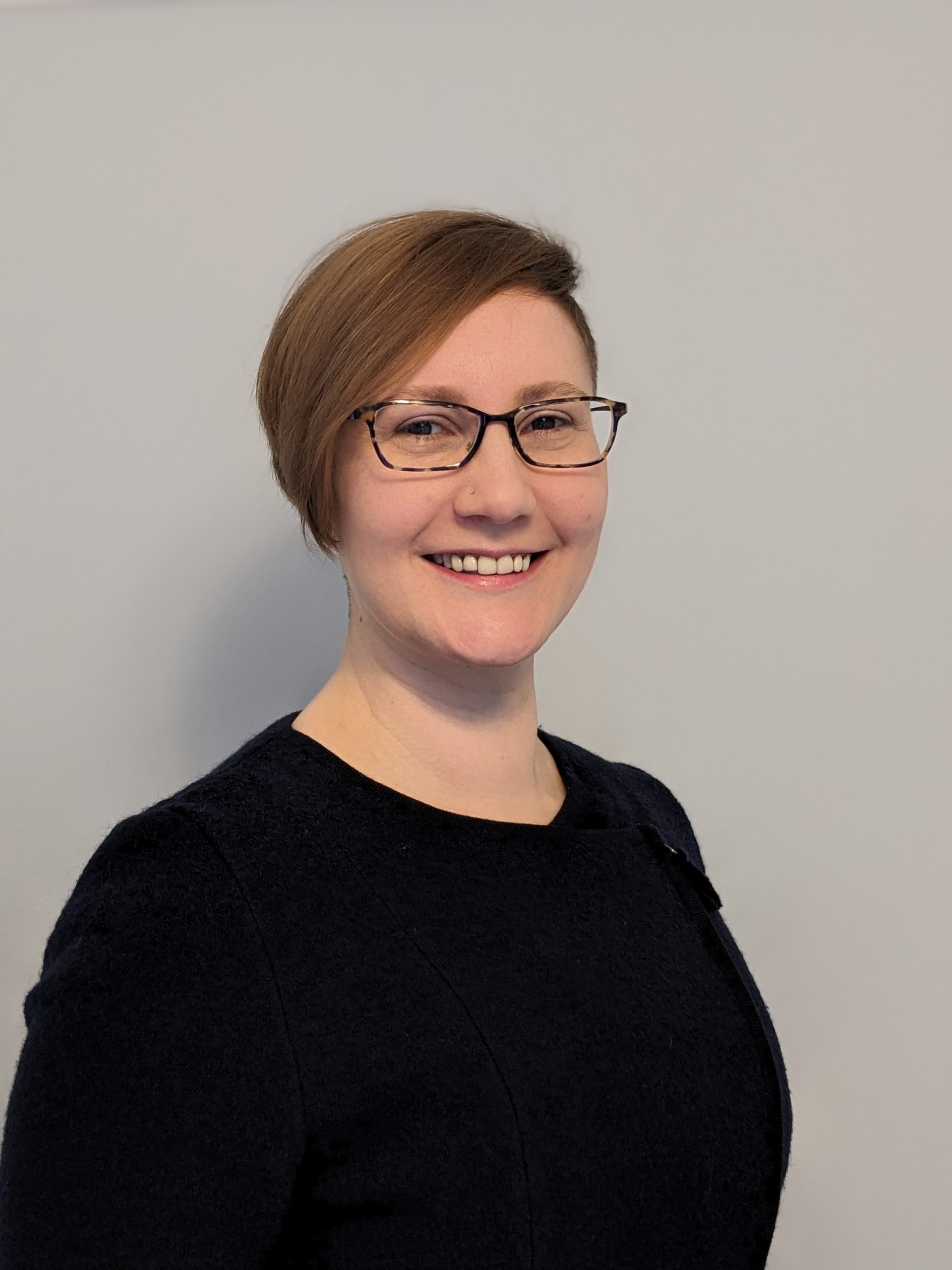 Kayla, a person with a short, blond, asymmetrical hairstyle, wears purple glasses and is smiling. 