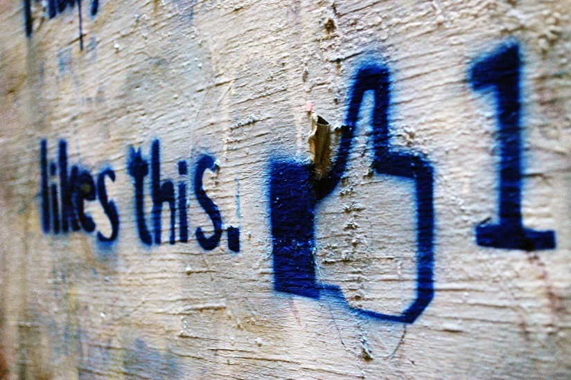 A graffiti style image with the words "likes this" and a thumbs up. It is spayed onto a white-washed wooden wall.