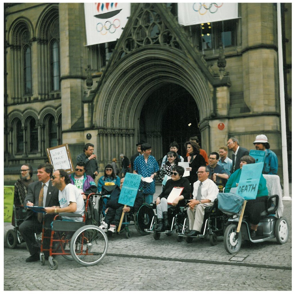 A group od GMCDP members speaking to a local MP in order to change the rights of disabled residents of Manchester. There are about 15 people either holding placards or paper. The MP is smiling and signing some papers infront of a camera