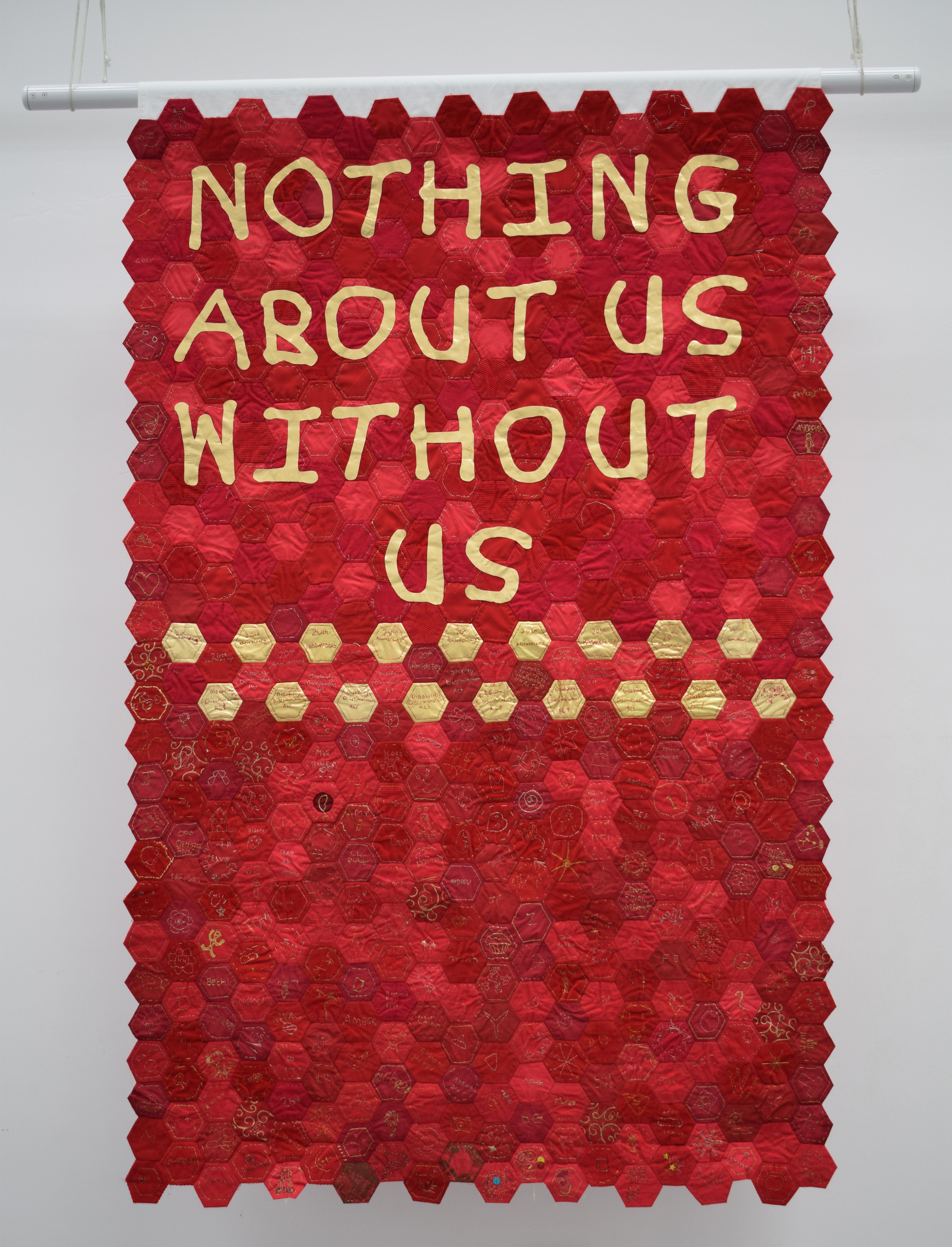A red banner made of hexagonal quilting fabric pieces with yellow-gold text “Nothing about us without us”. Beneath the text are two rows of hexagons alternating gold and red.