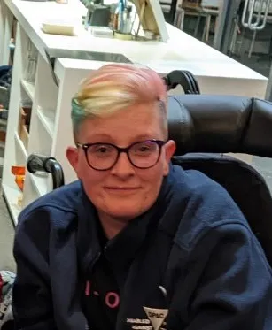 Photo shows Dennis Queen smiling, seated in a wheelchair. Dennis is white, she wears glasses and has short hair dyed in the colours of the trans pride flag