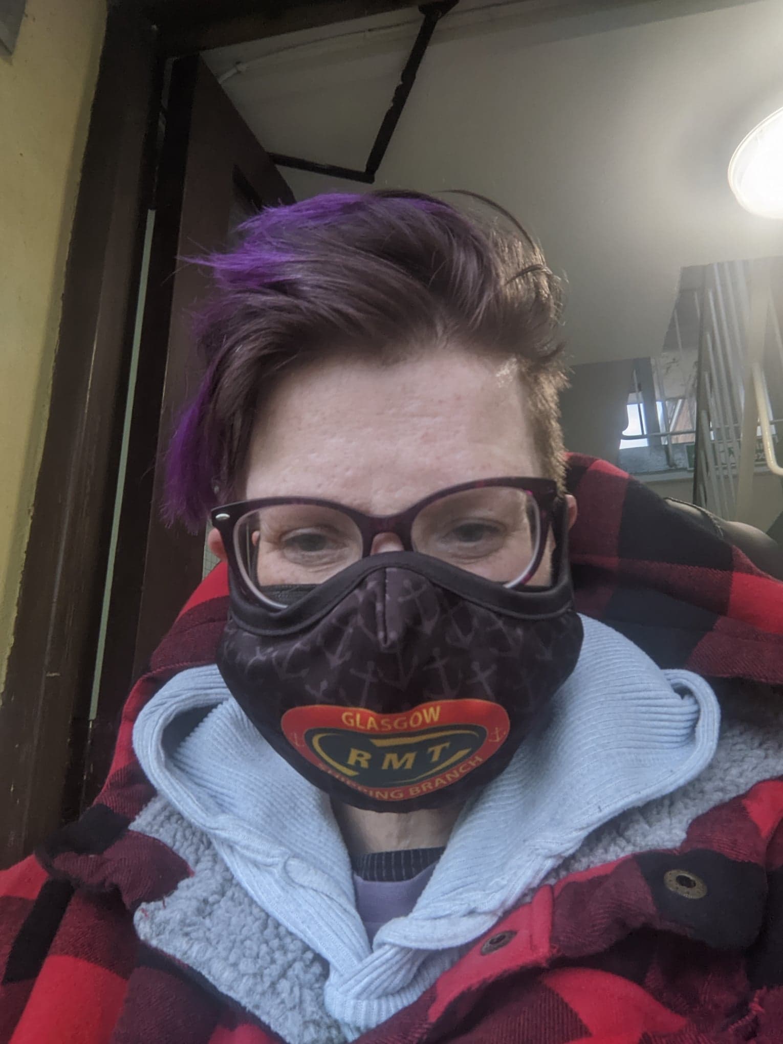 photograph of Dennis, a white person with dark purple-tipped hair wearing a hoodie and glasses, and an RMT face covering. The mask is black with dark grey anchors and in the middle a green and gold RMT logo. Text around the logo says "Glasgow Shipping Branch" in yellow on red