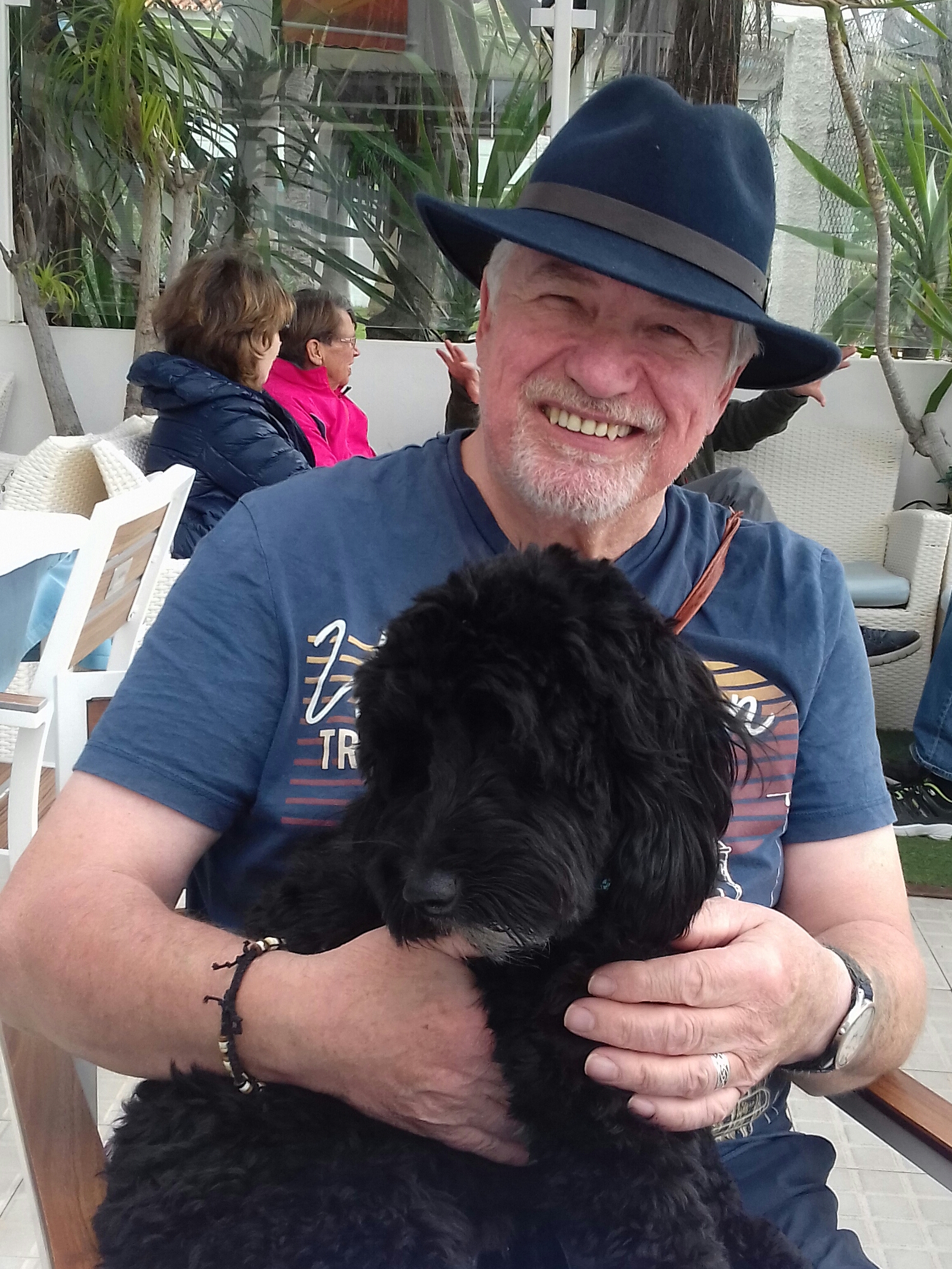Photograph shows Dave, of a white male sitting down with a black cockapoo dog on his lap. Dave is smiling and has a grey beard and moustache. He is wearing a blue fedora hat and a blue t-shirt.