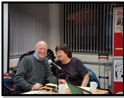 Joe Whittaker and Audrey Stanton sit next to each other by a microphone at a GMCDP event. They look like they have just finished speaking. They are both smiling at the camera.