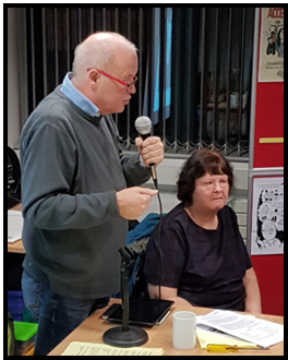 Photograph taken at a GMCDP event. Joe Whittaker is standing speaking into a microphone. Audrey Stanton sits beside him.