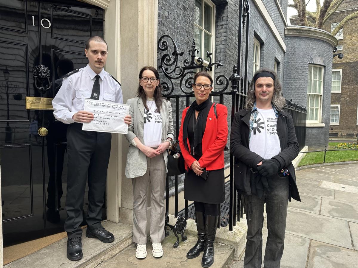 GMCDP board members Helen and Pete outside 10 Downing Street with MP Debbie Abrahams. The door is open and the guard holds a large envelope containing a petition letter from Greater Manchester Coalition of Disabled People which has just been handed in. Helen and Pete are wearing GMCDP t shirts. Debbie is wearing a red jacket.