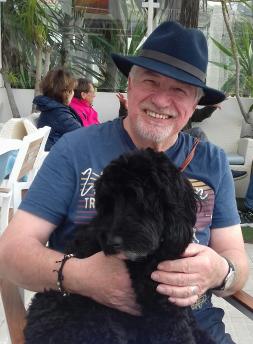 Photograph shows Dave, of a white male sitting down with a black cockapoo dog on his lap. Dave is smiling and has a grey beard and moustache. He is wearing a blue fedora hat and a blue t-shirt.