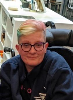 Photo shows Dennis Queen smiling, seated in a wheelchair. Dennis is white, she wears glasses and has short hair dyed in the colours of the trans pride flag