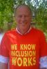 Photograph of Joe Whittaker, a white man in his 50s. He is outside in nature with a lake or river behind him and green leaves of a tree. He is facing the camera and smiling. He is wearing a red t-shirt with the slogan "we know inclusion works" in large bold capital lettering in yellow.